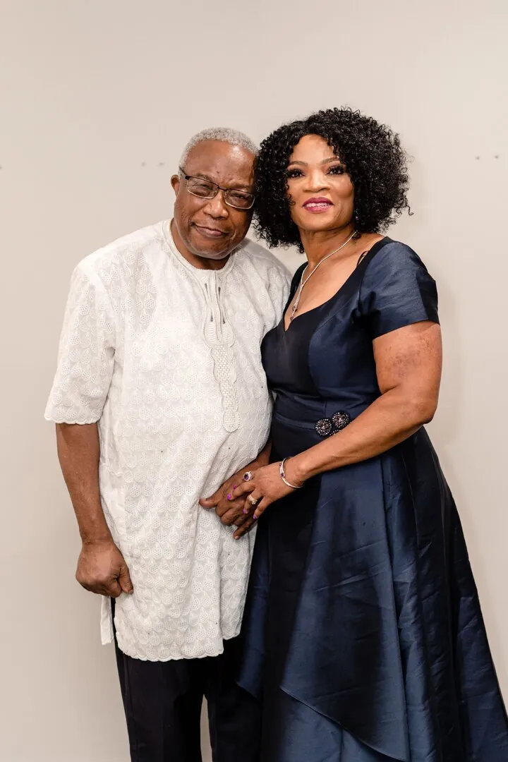 Nathan in a white lace-patterned tunic standing beside Ify in a navy satin dress, holding hands.