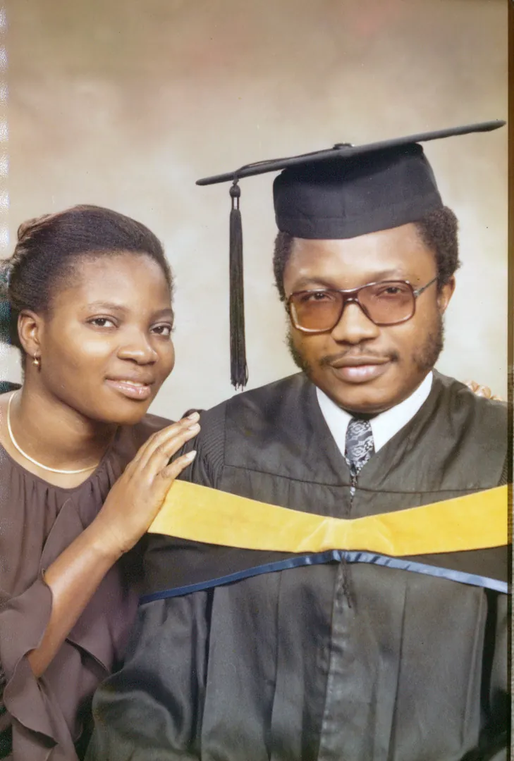 A young man in a graduation cap and gown, a woman beside him resting her hand on his shoulder.
