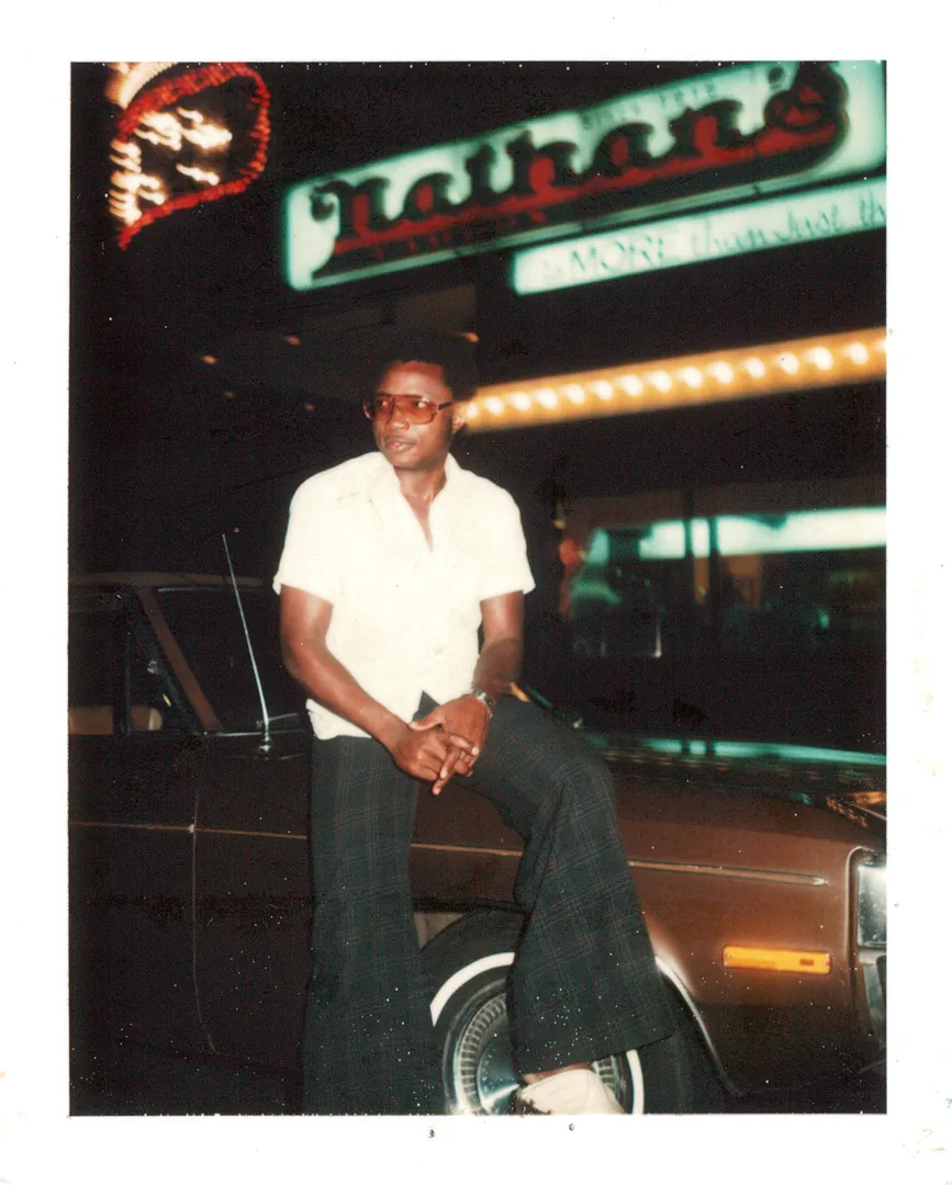 A young man in a white short-sleeved shirt and sunglasses sitting on a car at night beneath a neon sign.
