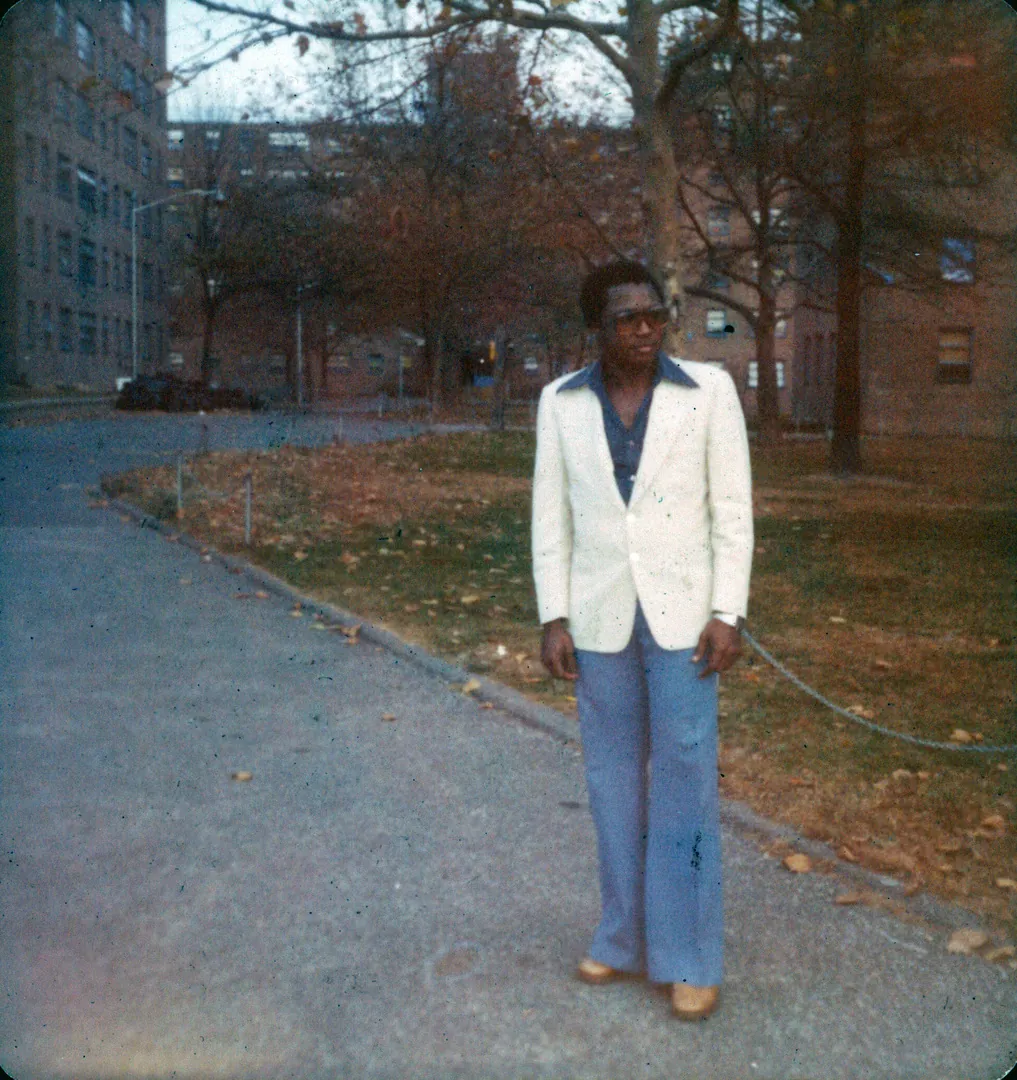 A young man in a white blazer and flared blue trousers standing on a path between apartment buildings in New York.