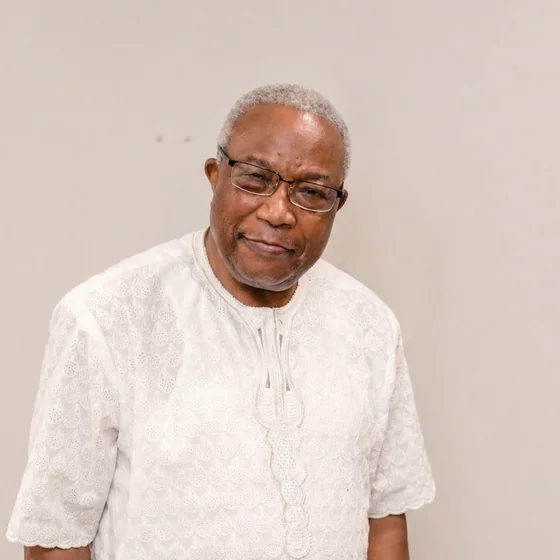 Nathan Momah in a white embroidered tunic, silver hair and glasses, smiling warmly with his head tilted slightly.
