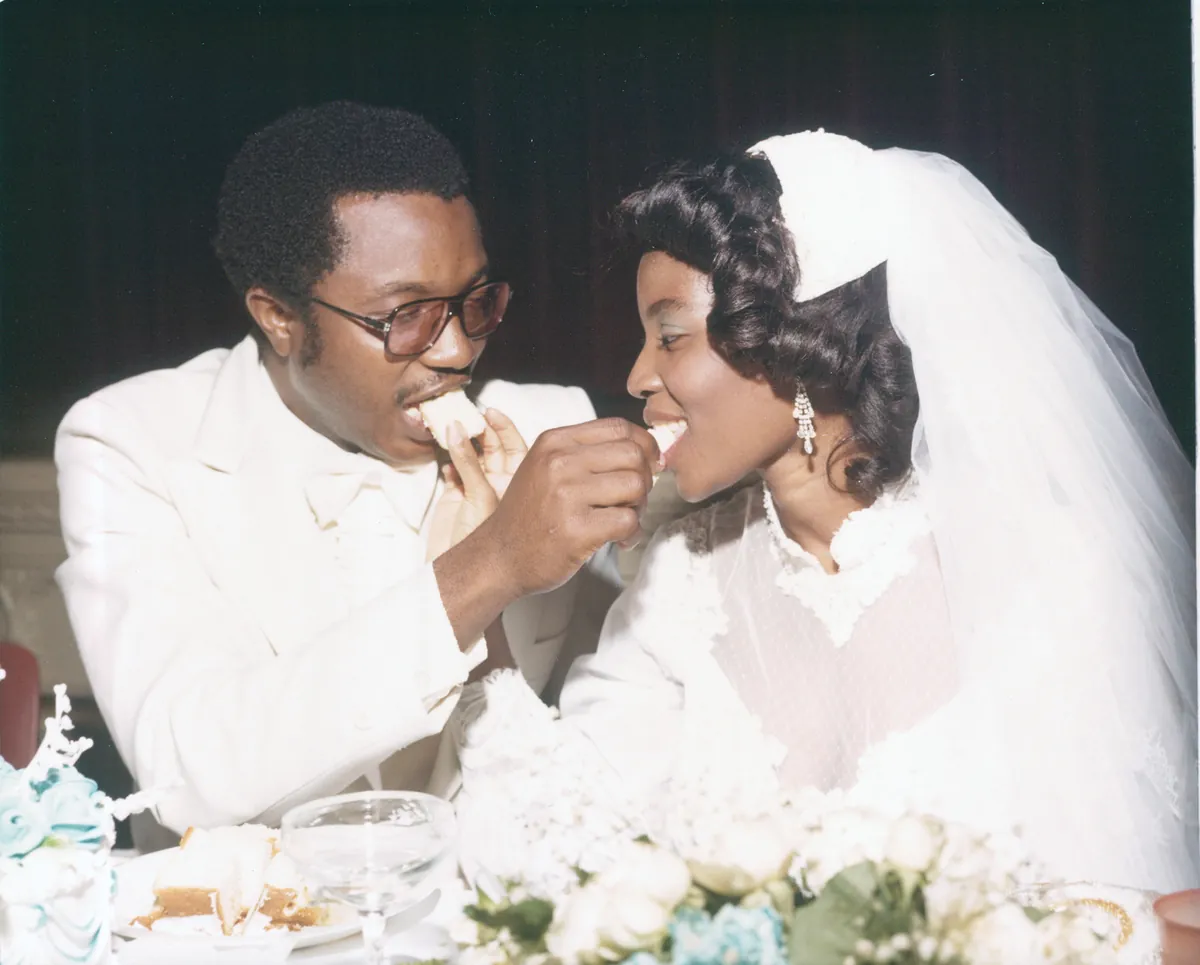 A groom in a white tuxedo feeding wedding cake to his bride in her veil.