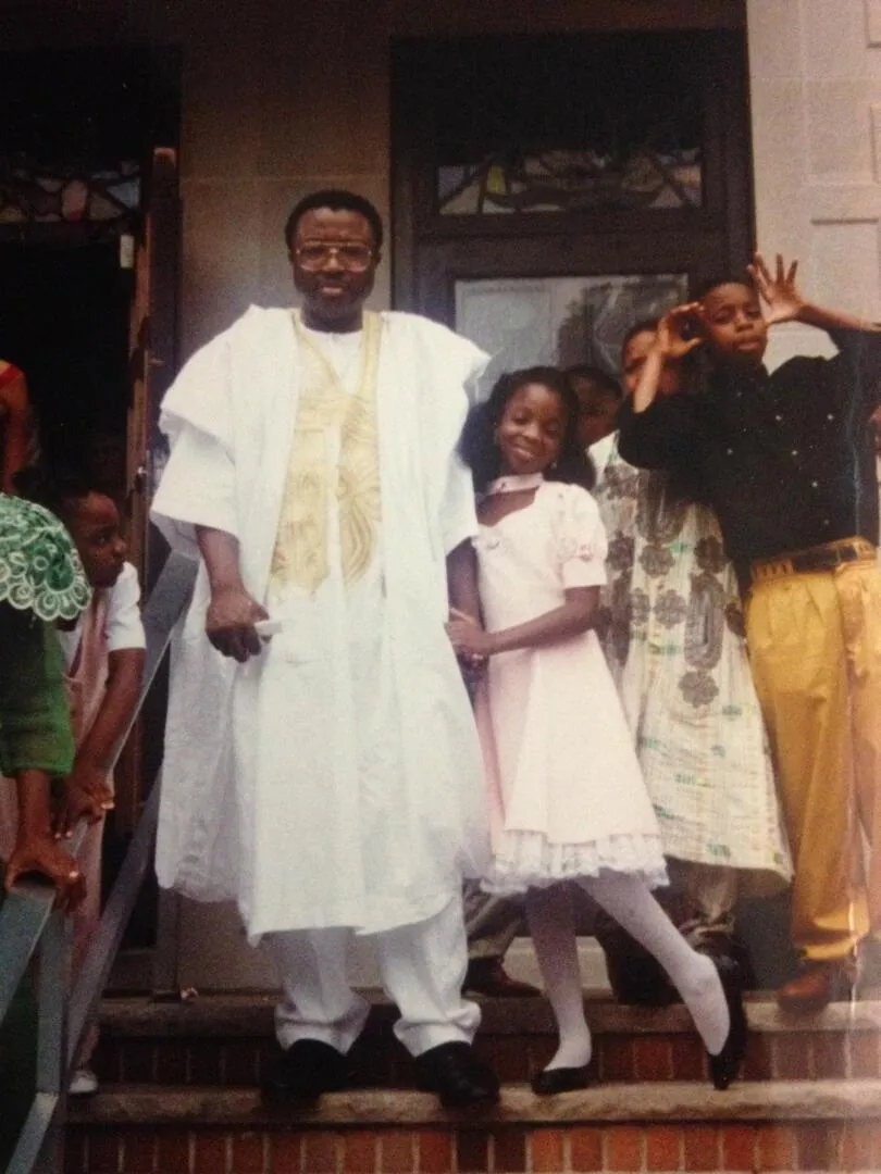 Nathan in a white agbada with gold embroidery standing on porch steps, two girls in white dresses beside him.